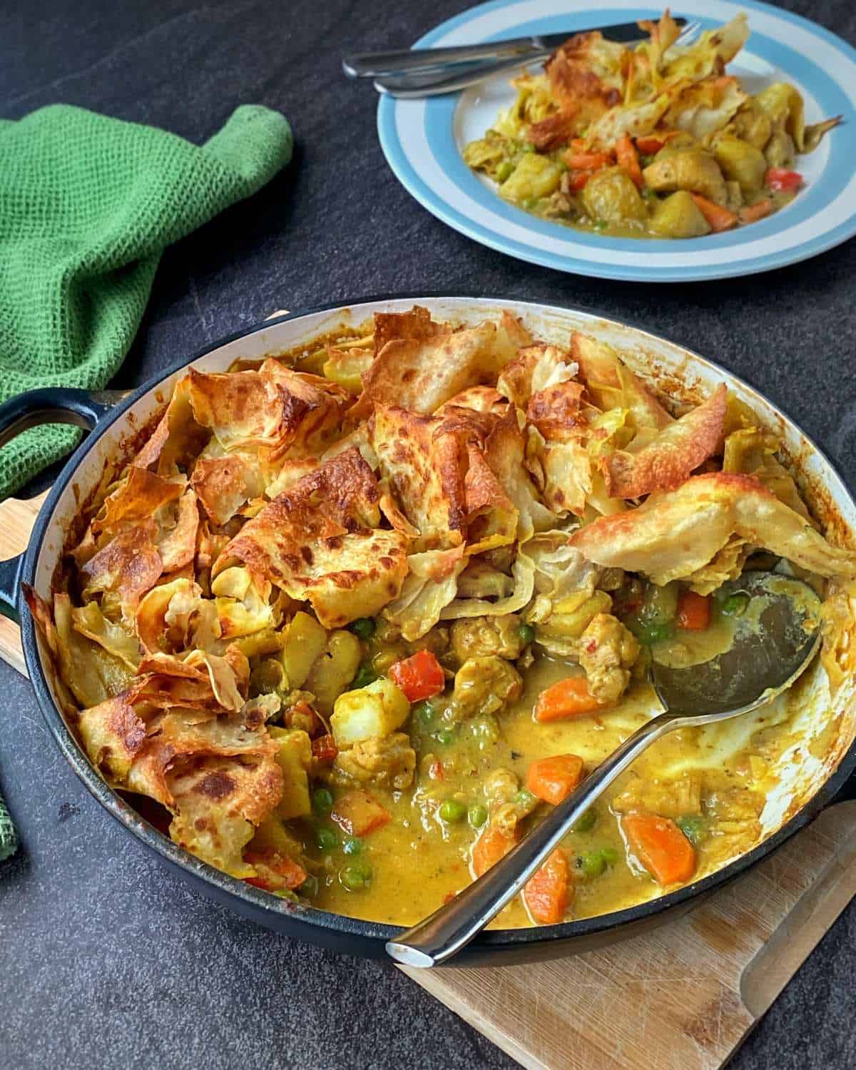 Chicken Curry Roti Pie A casserole dish filled with a vegetable pot pie topped with crispy pastry, next to a plate of a serving. A green towel is placed nearby on a dark countertop.