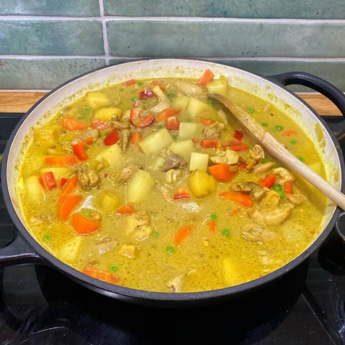 Chicken Curry Roti Pie A pot of yellow curry with chicken, potatoes, carrots, red bell peppers, peas, and a wooden spoon on the side, simmering on a stovetop.