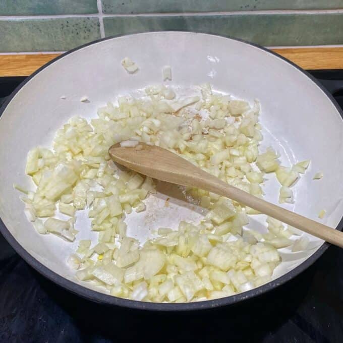 Chicken Curry Roti Pie Chopped onions cooking in a white pan with a wooden spoon resting inside.