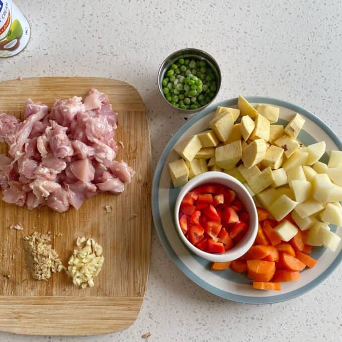 Chicken Curry Roti Pie Chopped raw chicken, minced garlic and ginger on a wooden board next to a plate with chopped potatoes, sweet potatoes, carrots, red pepper, and a small bowl of green peas.