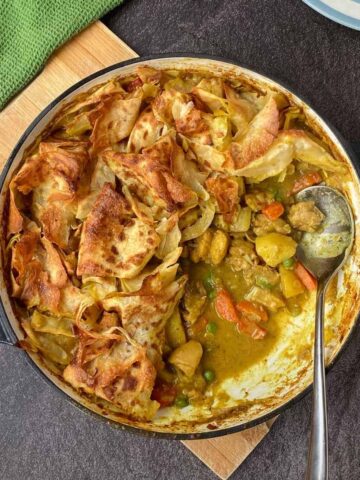 A round baking dish filled with a vegetable pot pie topped with golden, flaky pastry, partially served with a metal spoon.