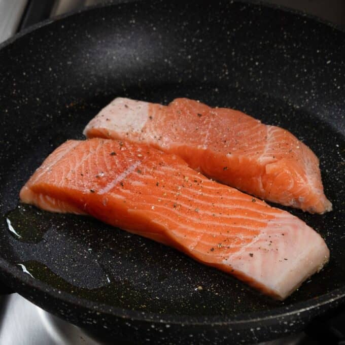 Salmon, Asparagus Piccata Pasta Two raw salmon fillets with seasoning are placed in a black frying pan with a small amount of oil.