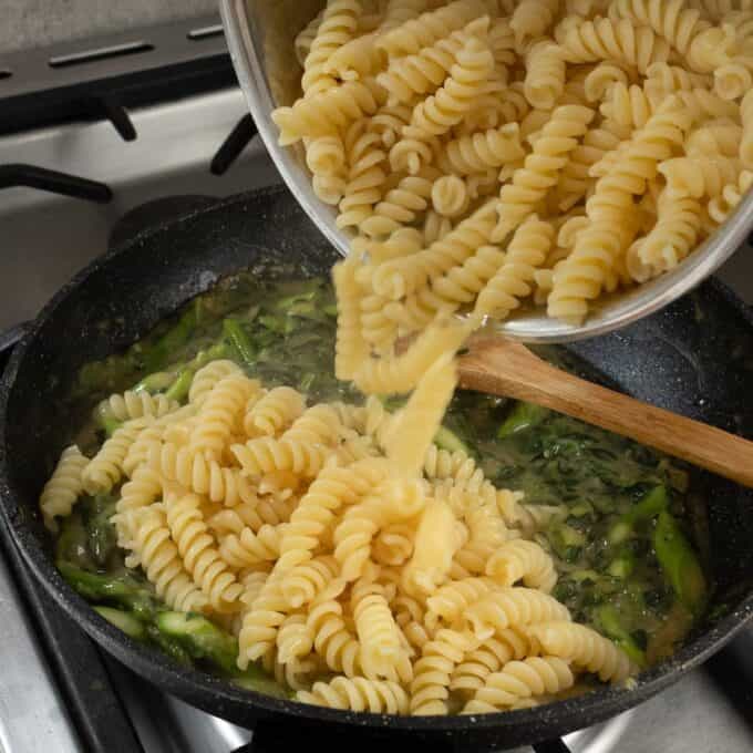Salmon, Asparagus Piccata Pasta Cooked rotini pasta is being poured from a pot into a pan with green vegetables and sauce on a stove.