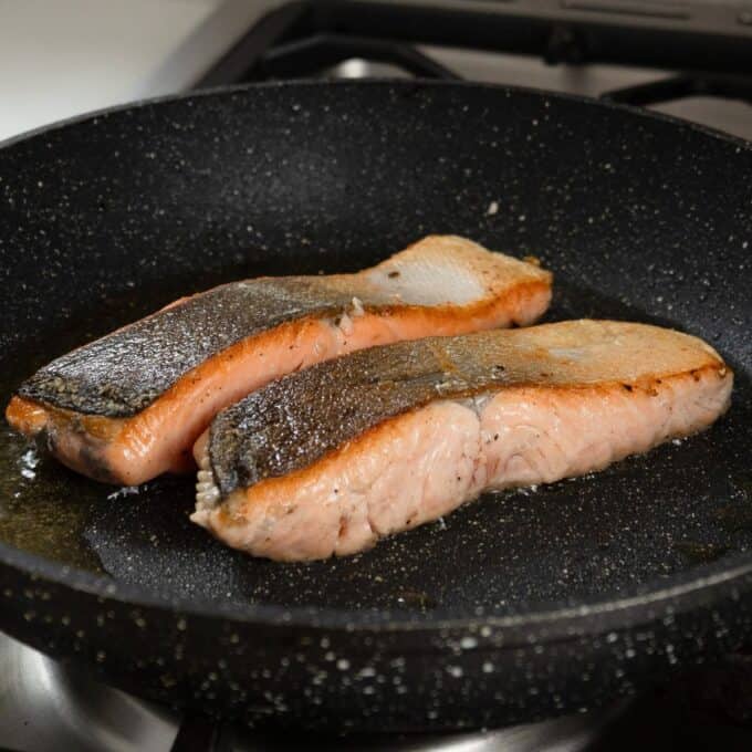 Salmon, Asparagus Piccata Pasta Two salmon fillets with skin are being pan-seared in a black nonstick frying pan on a stovetop.