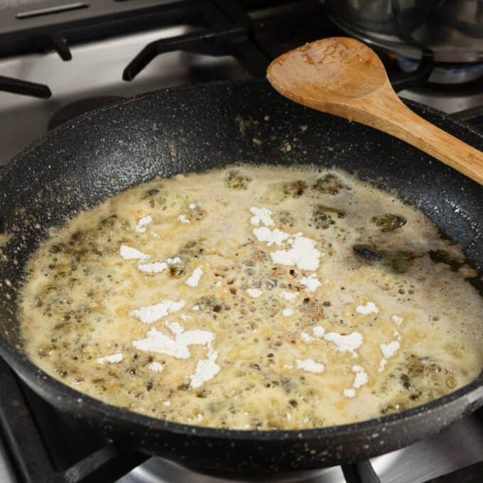 Salmon, Asparagus Piccata Pasta A frying pan on a stove contains bubbling brown liquid with some flour sprinkled on top and a wooden spoon resting on the edge.