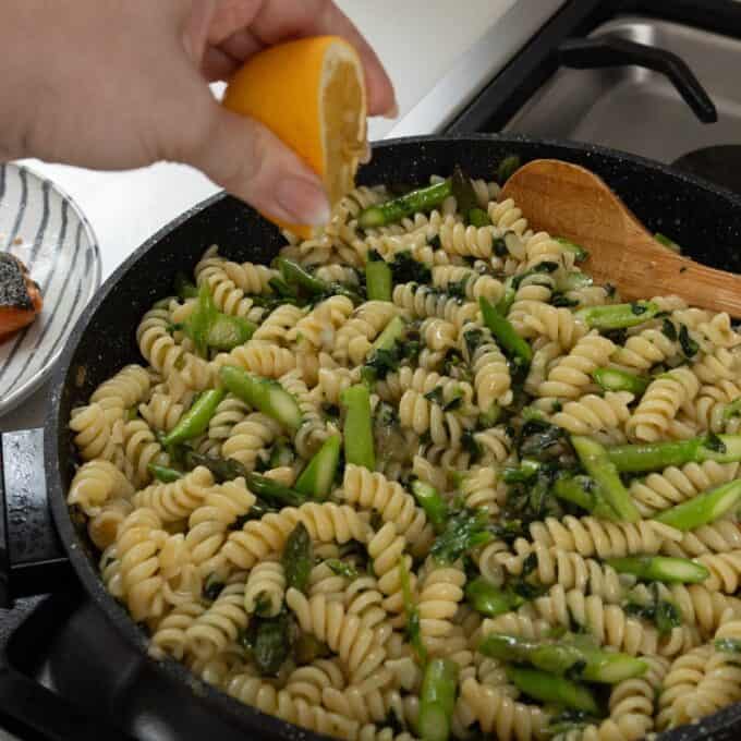 Salmon, Asparagus Piccata Pasta A hand squeezes a lemon over a pan of cooked rotini pasta with asparagus and greens, being stirred with a wooden spoon on a stovetop.