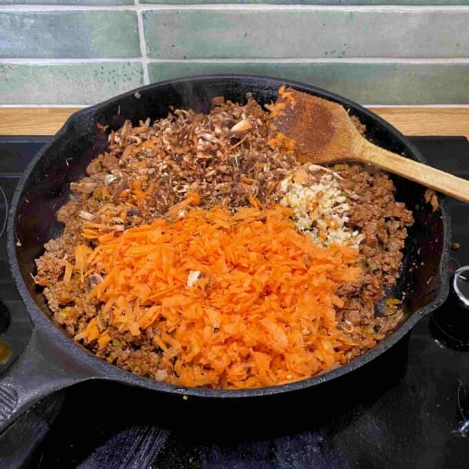Beef Mince Pie with Kumara Mash A cast iron skillet on a stove contains ground meat, grated carrot, chopped mushrooms, and minced garlic, being cooked and mixed with a wooden spoon.