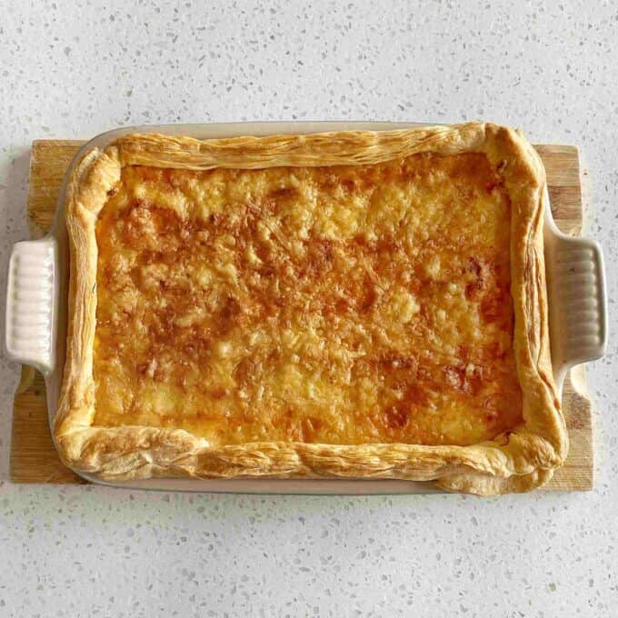 Beef Mince Pie with Kumara Mash A rectangular baking dish filled with a golden-brown baked pie topped with melted cheese, set on a wooden board.