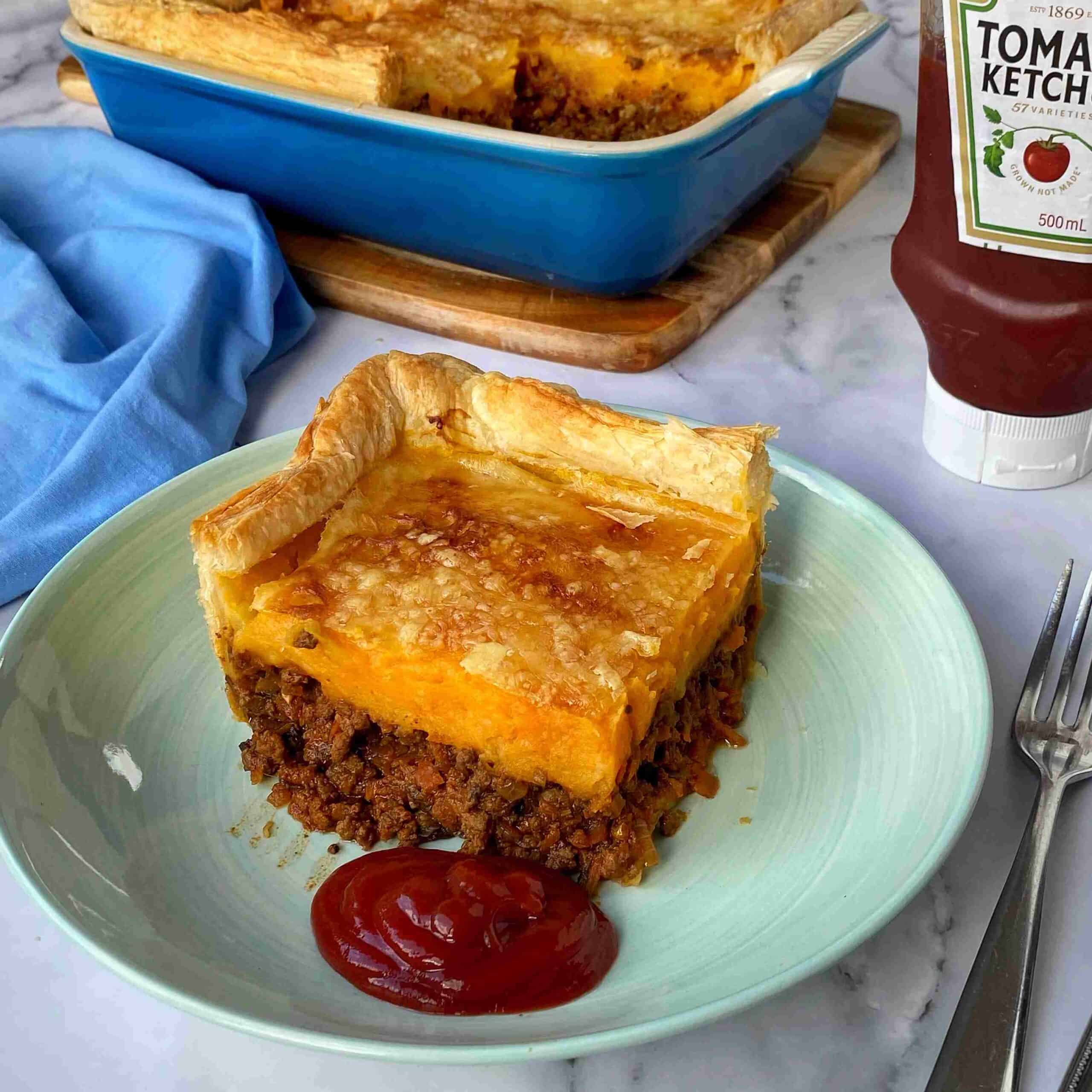 Beef Mince Pie with Kumara Mash A slice of meat pie topped with pastry and served with ketchup on a plate; the remaining pie and a bottle of ketchup are in the background.