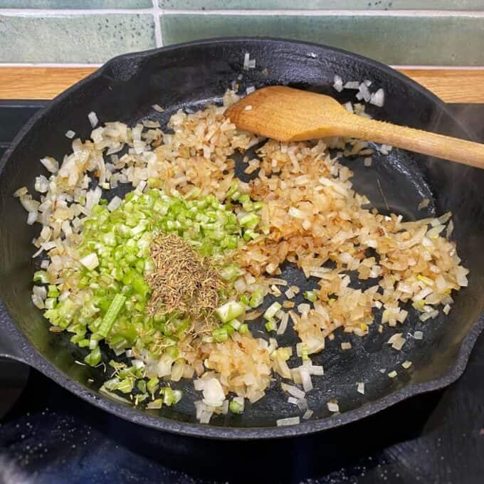 Beef Mince Pie with Kumara Mash Chopped onions, celery, and dried herbs are being sautéed in a cast iron skillet with a wooden spoon.