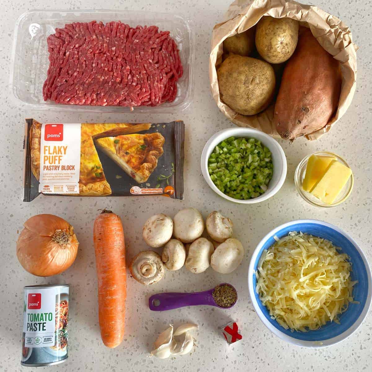 Beef Mince Pie with Kumara Mash Assorted ingredients on a countertop, including ground beef, potatoes, sweet potato, puff pastry, chopped celery, butter, onion, carrot, mushrooms, shredded cheese, tomato paste, and garlic cloves.