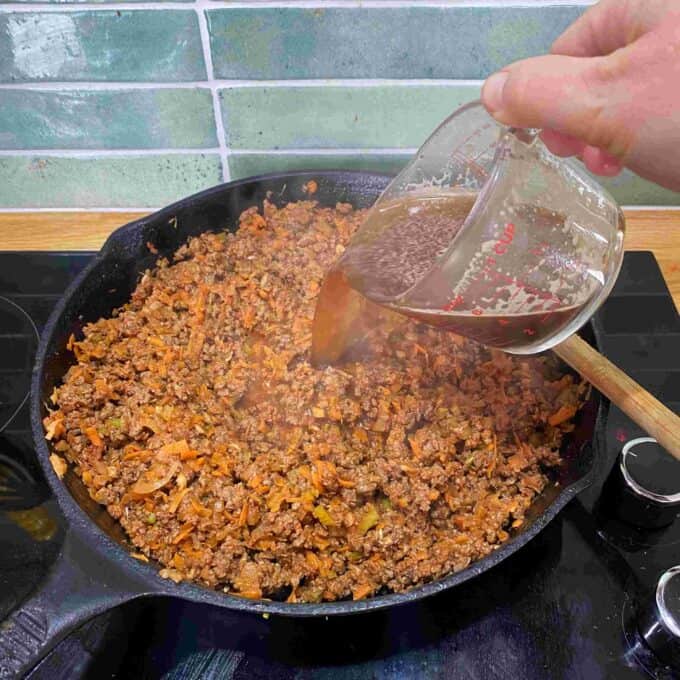 Beef Mince Pie with Kumara Mash A hand pours liquid from a measuring jug into a skillet filled with minced meat and vegetables on a stovetop.