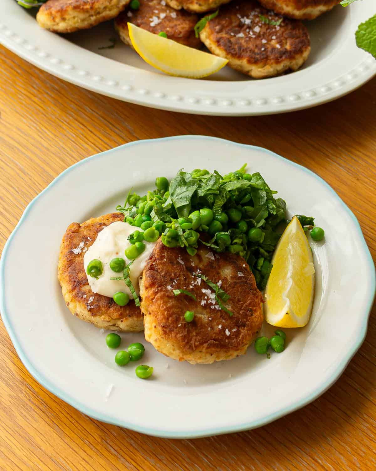 Two golden fish cakes served with green peas, a dollop of mayonnaise, fresh greens, and a lemon wedge on a white plate.