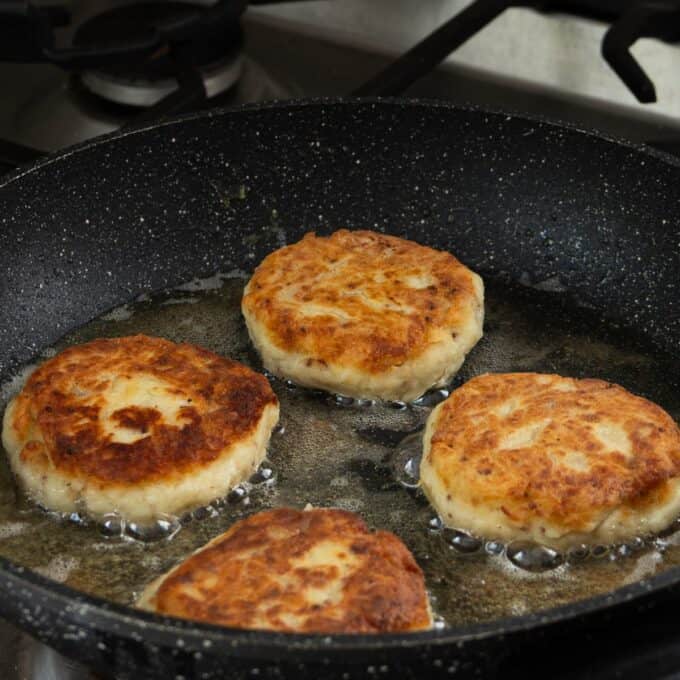 Four golden-brown patties frying in oil on a black skillet, placed on a stovetop.
