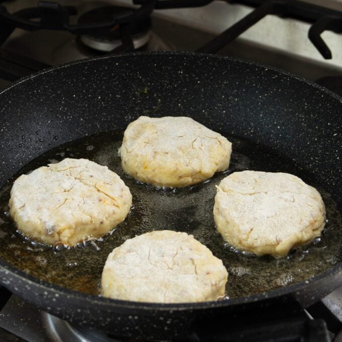 Four uncooked patties are being fried in oil in a black skillet on a stovetop.