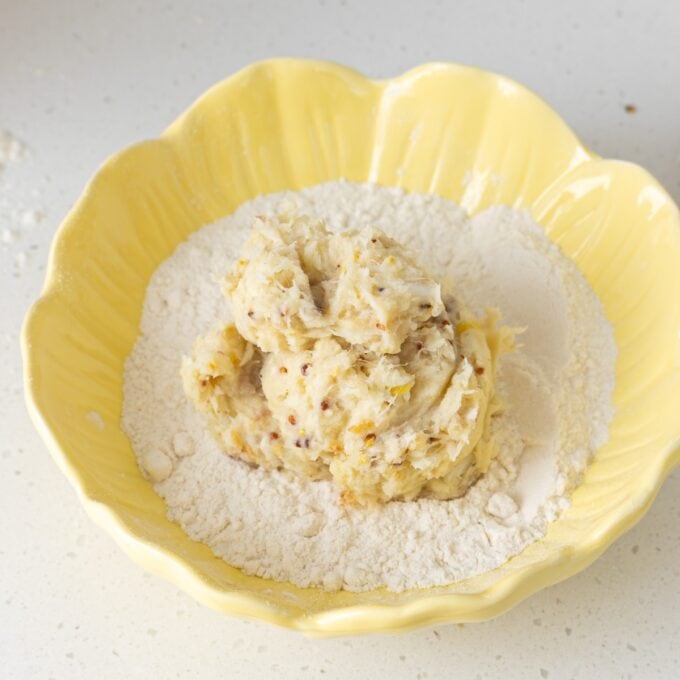 A yellow bowl containing a mound of dough placed on top of loose flour on a light-colored countertop.