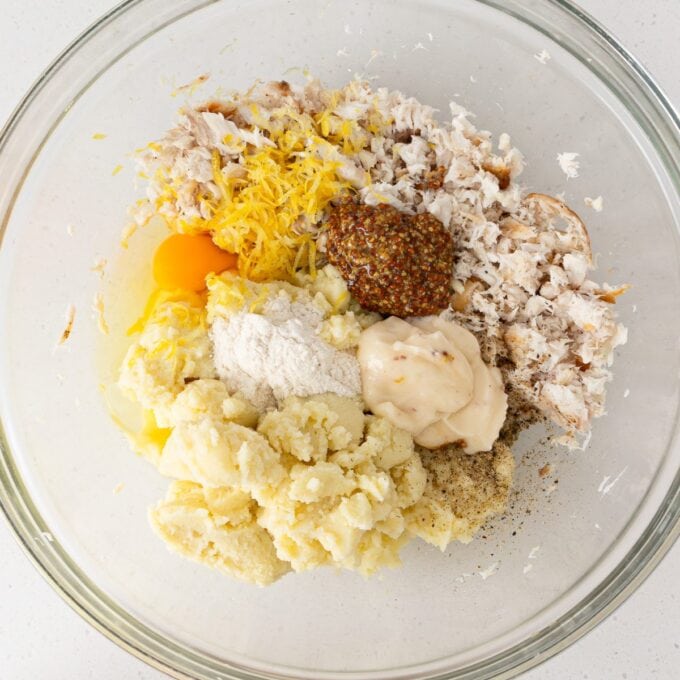 A glass bowl containing crab meat, egg, mashed potatoes, lemon zest, whole grain mustard, mayonnaise, flour, salt, and pepper, ready to be mixed.