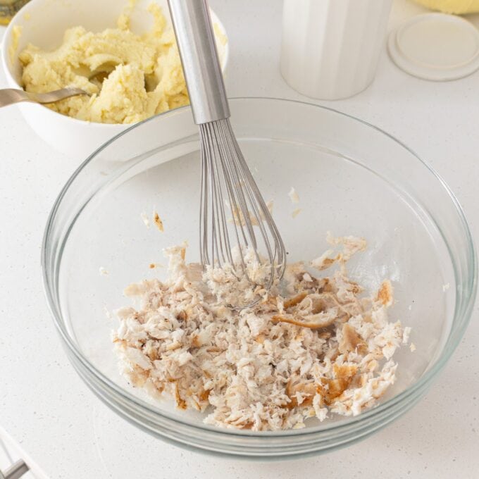 A large glass bowl with shredded cooked chicken being mixed with a whisk; a bowl of mashed potatoes is in the background on a white countertop.