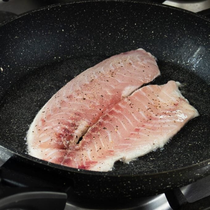 Two raw fish fillets seasoned with pepper are being cooked in a black frying pan on a stovetop.