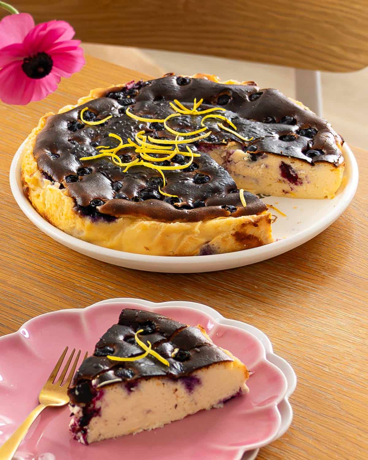 Lemon Blueberry Basque Cheesecake A sliced cheesecake topped with blueberries and lemon zest sits on a plate, with one piece served on a pink dish beside a gold fork.