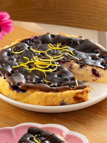 A partially sliced cheesecake topped with blueberries and lemon zest sits on a white plate on a wooden table.