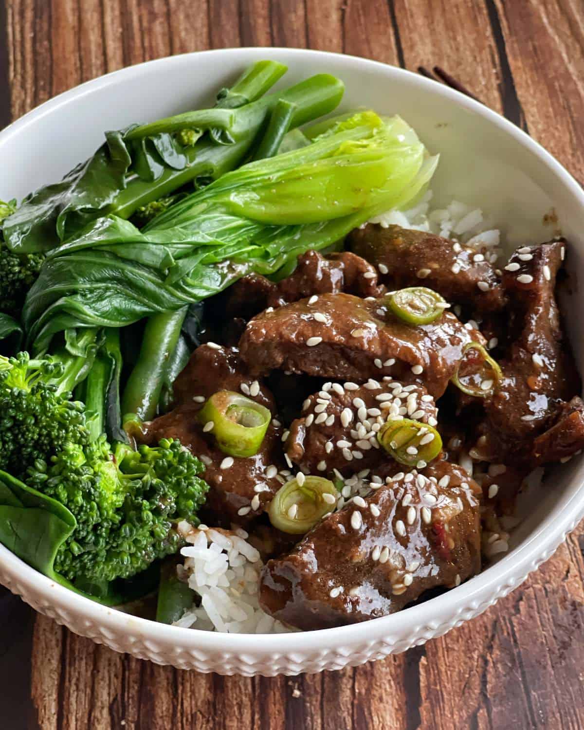 A bowl of white rice topped with cooked beef in sauce, garnished with sesame seeds and green onions, served with steamed greens and broccoli.
