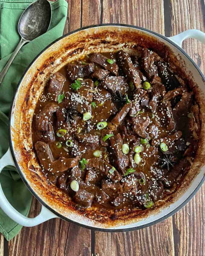 5 Spice Beef A pot of braised beef in dark sauce topped with sliced green onions and sesame seeds, with a serving spoon and green napkin beside it on a wooden surface.