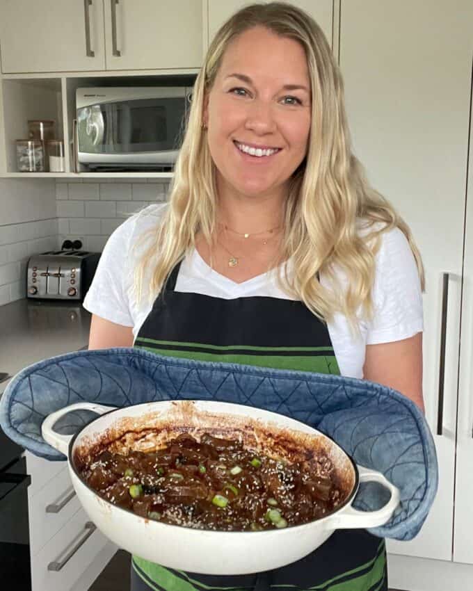 5 Spice Beef A woman in a kitchen holding a round white dish with a cooked stew, wearing a striped apron and oven mitts, and smiling at the camera.