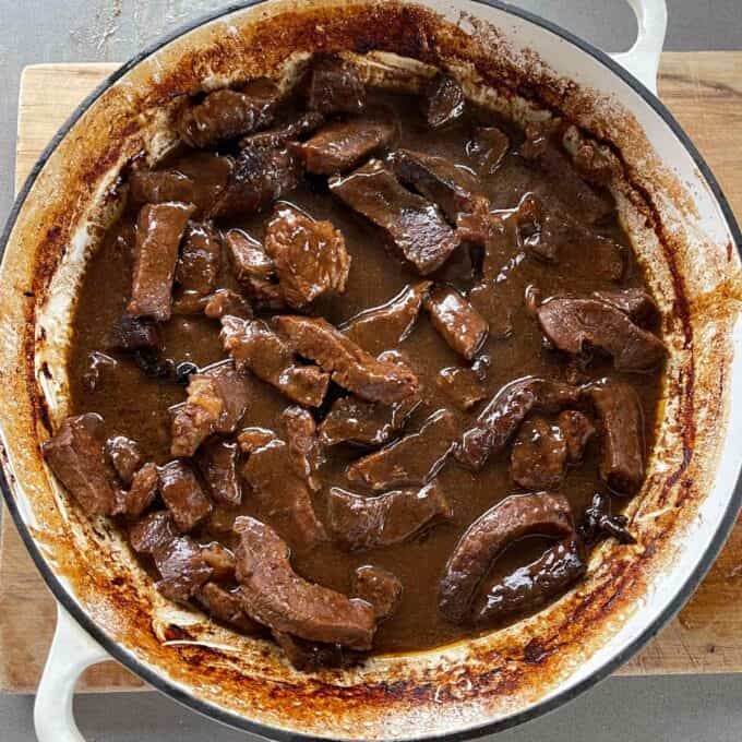 5 Spice Beef Chunks of beef simmering in a thick brown gravy inside a round white enamel pot on a wooden surface.