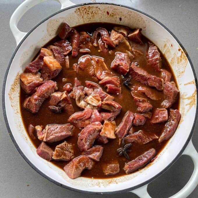 5 Spice Beef Chunks of raw beef marinating in a brown sauce with visible spices, inside a white round pot on a gray surface.