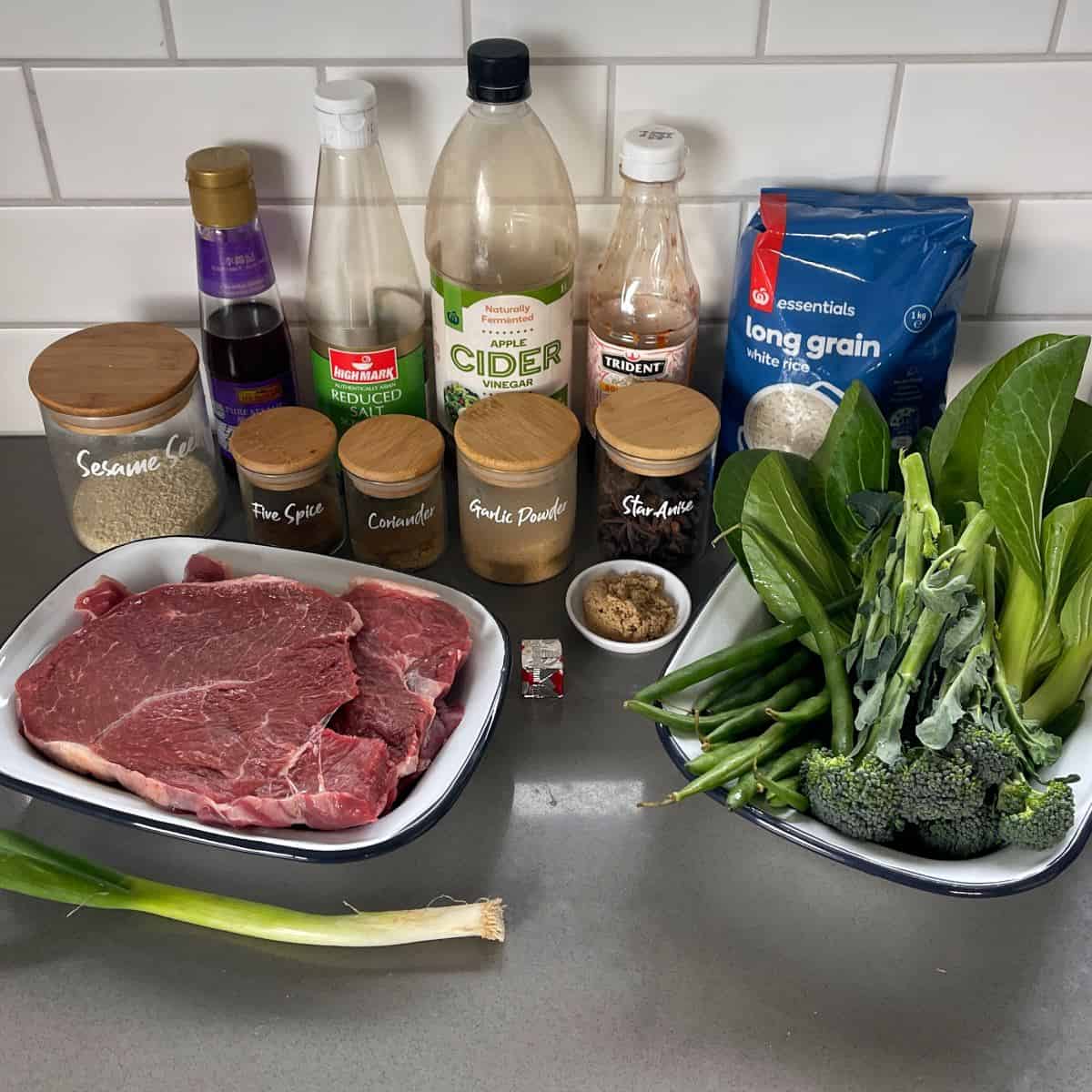 5 Spice Beef Raw beef steaks, fresh green vegetables, various sauces, spices, and a bag of long grain white rice arranged on a kitchen counter.