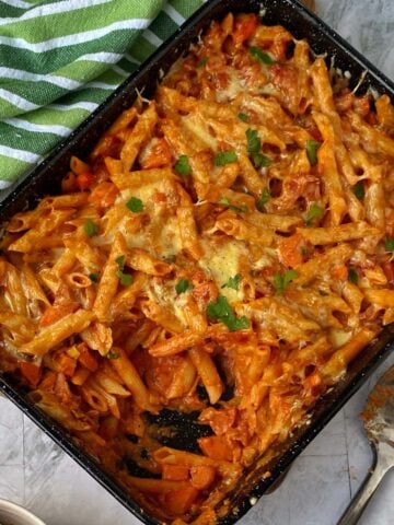 A baking dish filled with Bacon Veggie Pasta Bake in a rich tomato sauce, topped with melted cheese and garnished with parsley. A serving spoon rests beside the dish.