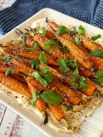 A plate of roasted carrots garnished with fresh mint leaves, served on a layer of creamy white sauce, on a white square dish.
