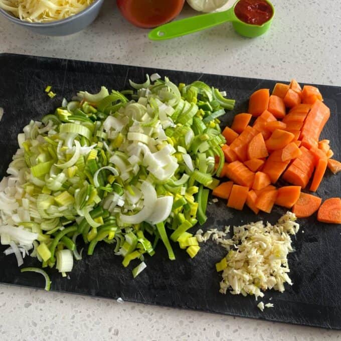 Bacon Veggie Pasta Bake Chopped leeks, diced carrots, and minced garlic arranged on a black cutting board, with bowls and measuring cups in the background.