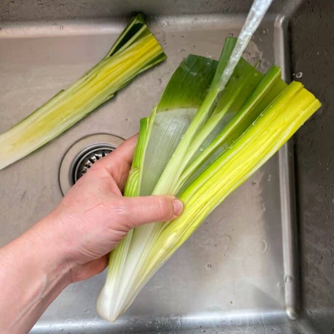Bacon Veggie Pasta Bake A hand holds a leek under running water in a sink, cleaning it; another leek is lying in the sink.