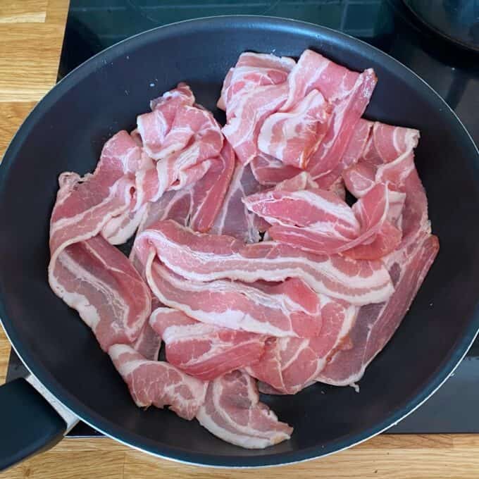 Bacon Veggie Pasta Bake Raw bacon strips arranged in a single layer inside a black frying pan on a stovetop, ready to be cooked.