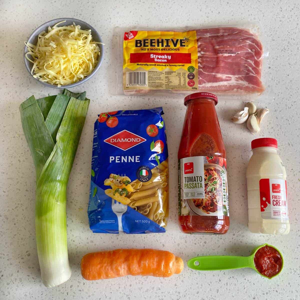 Bacon Veggie Pasta Bake Ingredients for a pasta dish laid out on a countertop, including penne, leek, carrot, bacon, tomato passata, cream, cheese, garlic, and a small bowl of oil.