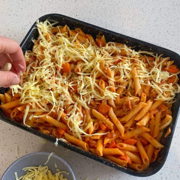 Bacon Veggie Pasta Bake A hand sprinkles shredded cheese over a tray of penne pasta mixed with tomato sauce and vegetables on a kitchen countertop.