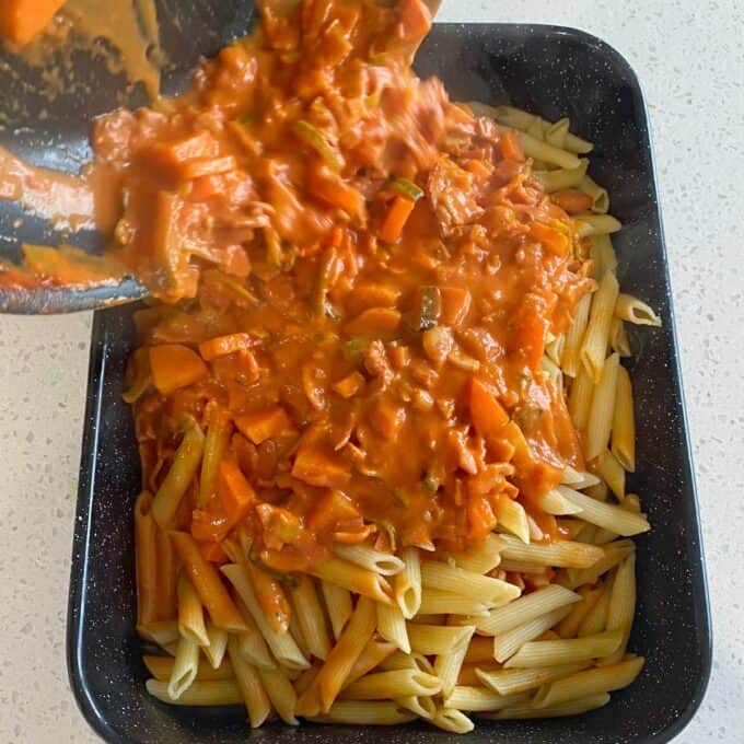 Bacon Veggie Pasta Bake Cooked penne pasta in a black tray is being topped with a tomato-based vegetable sauce from a pan.