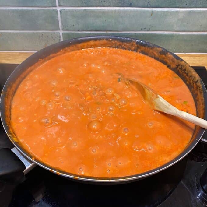 Bacon Veggie Pasta Bake A pan of tomato sauce is simmering on a stovetop, with bubbles forming and a wooden spoon resting inside.