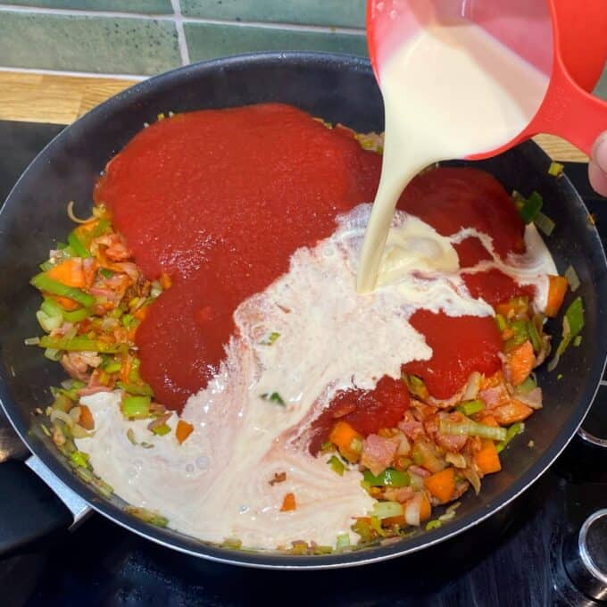 Bacon Veggie Pasta Bake A hand pours cream from a measuring cup into a frying pan containing tomato sauce, chopped vegetables, and bacon on a stovetop.