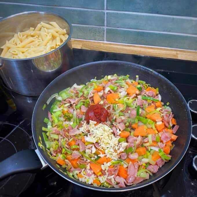 Bacon Veggie Pasta Bake A pan with chopped vegetables, bacon, tomato paste, and minced garlic cooking on a stove, with a pot of cooked penne pasta in the background.