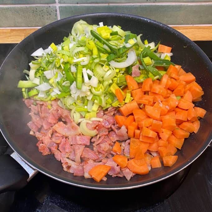 Bacon Veggie Pasta Bake A frying pan containing chopped bacon, sliced leeks, and diced carrots, arranged in sections, on a stovetop.