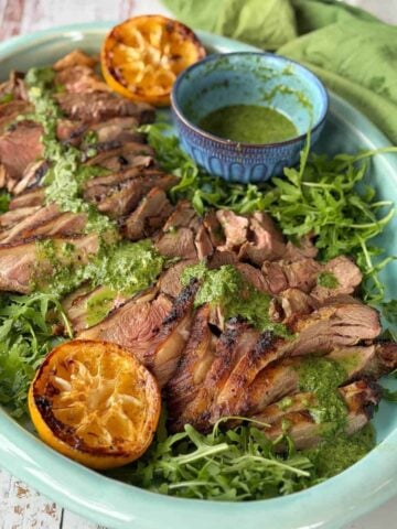 Sliced grilled lamb on a bed of arugula with green sauce drizzled on top, served with grilled lemon halves and a bowl of sauce on a large platter.