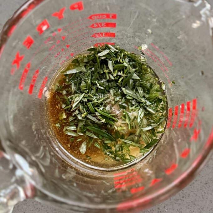 BBQ Lamb with Fresh Mint Sauce A glass measuring cup containing chopped fresh herbs and a brown liquid mixture on a gray surface.