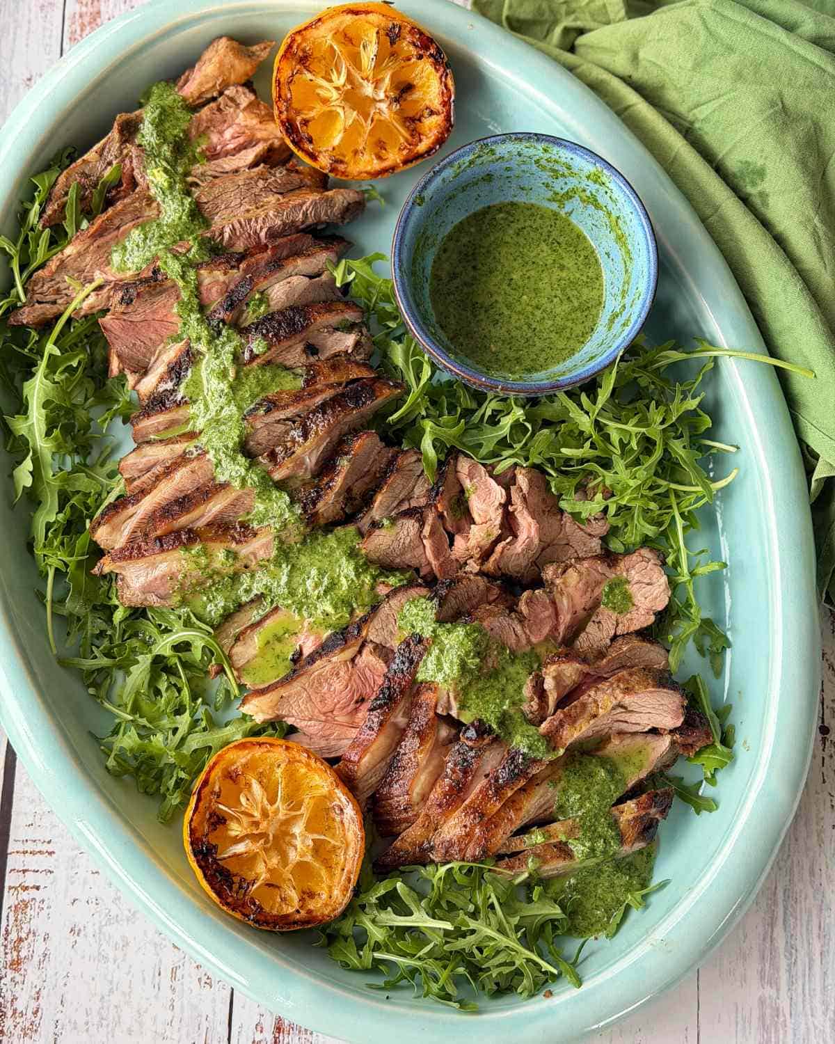 BBQ Lamb with Fresh Mint Sauce Sliced grilled steak on a bed of arugula, topped with green sauce, served with grilled lemon halves and a small bowl of extra sauce on a light blue platter.