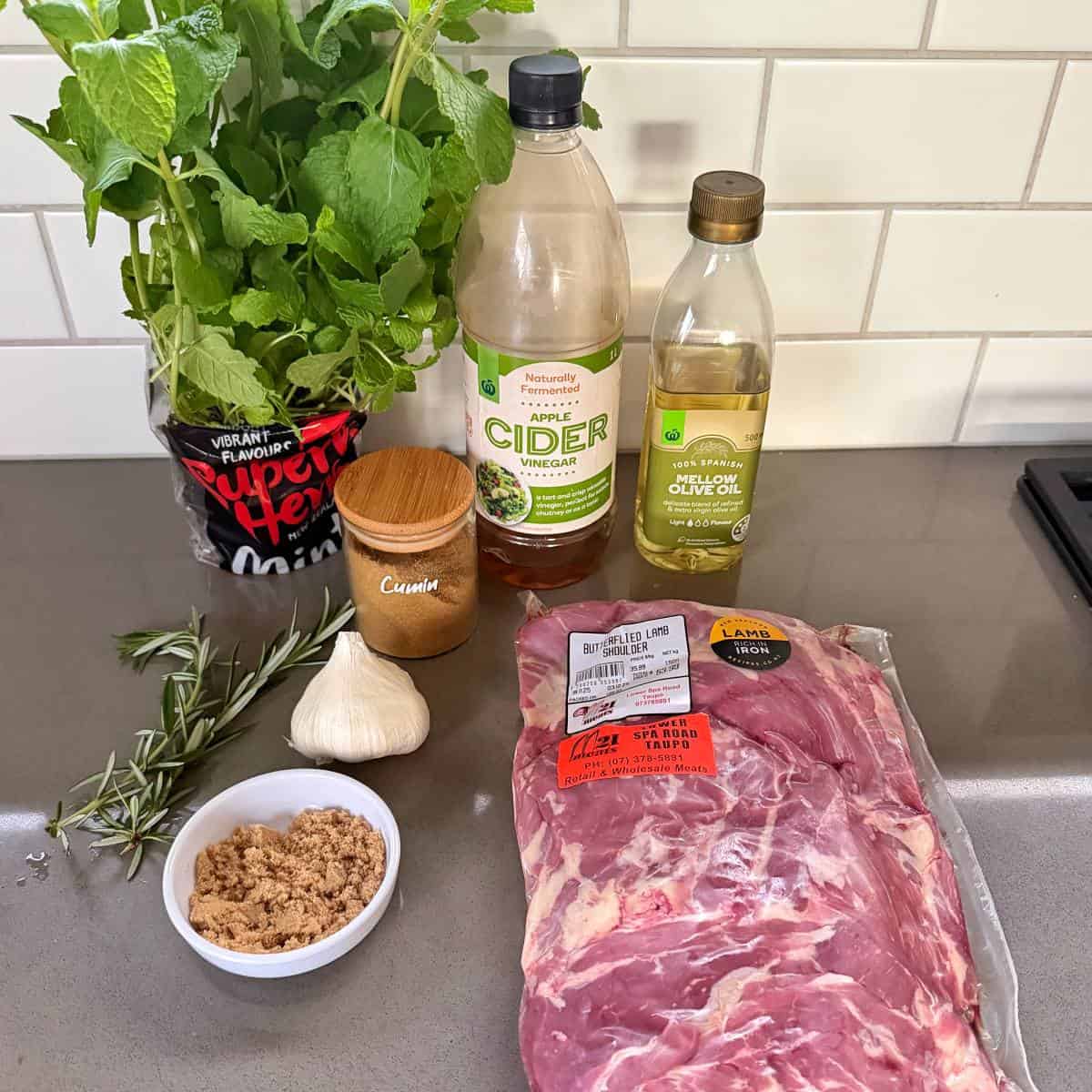 BBQ Lamb with Fresh Mint Sauce A package of spareribs, brown sugar, garlic, rosemary, mint, cumin, apple cider vinegar, and olive oil arranged on a kitchen counter.