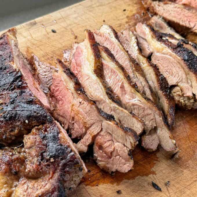 BBQ Lamb with Fresh Mint Sauce Sliced grilled steak on a wooden cutting board, showing a medium rare interior with a seared, charred crust.