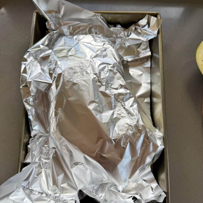 BBQ Lamb with Fresh Mint Sauce A metal baking tray with food covered by crumpled aluminum foil, resting on a gray countertop.