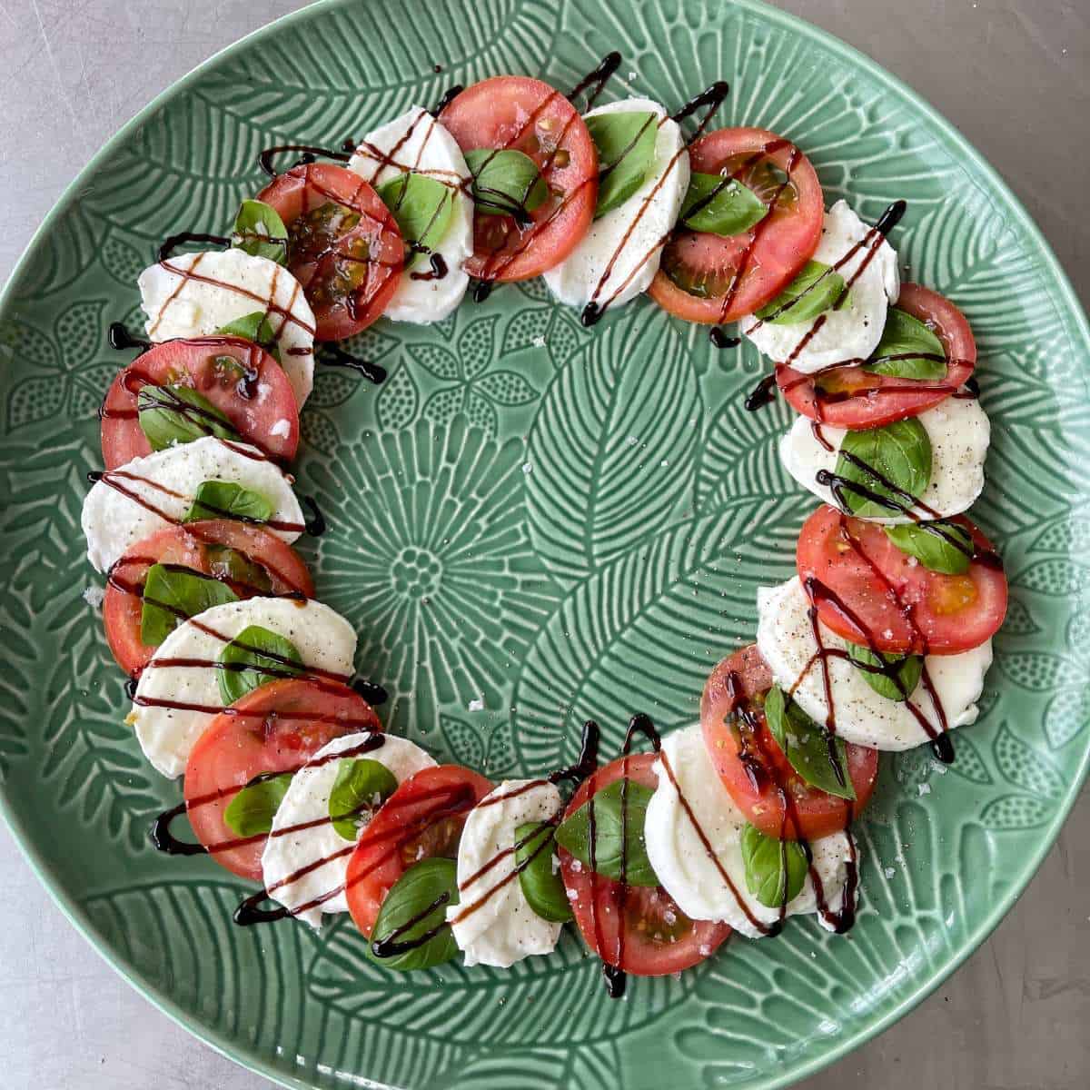 Caprese Wreath A circular arrangement of tomato, mozzarella, and basil slices on a green plate, drizzled with balsamic glaze.